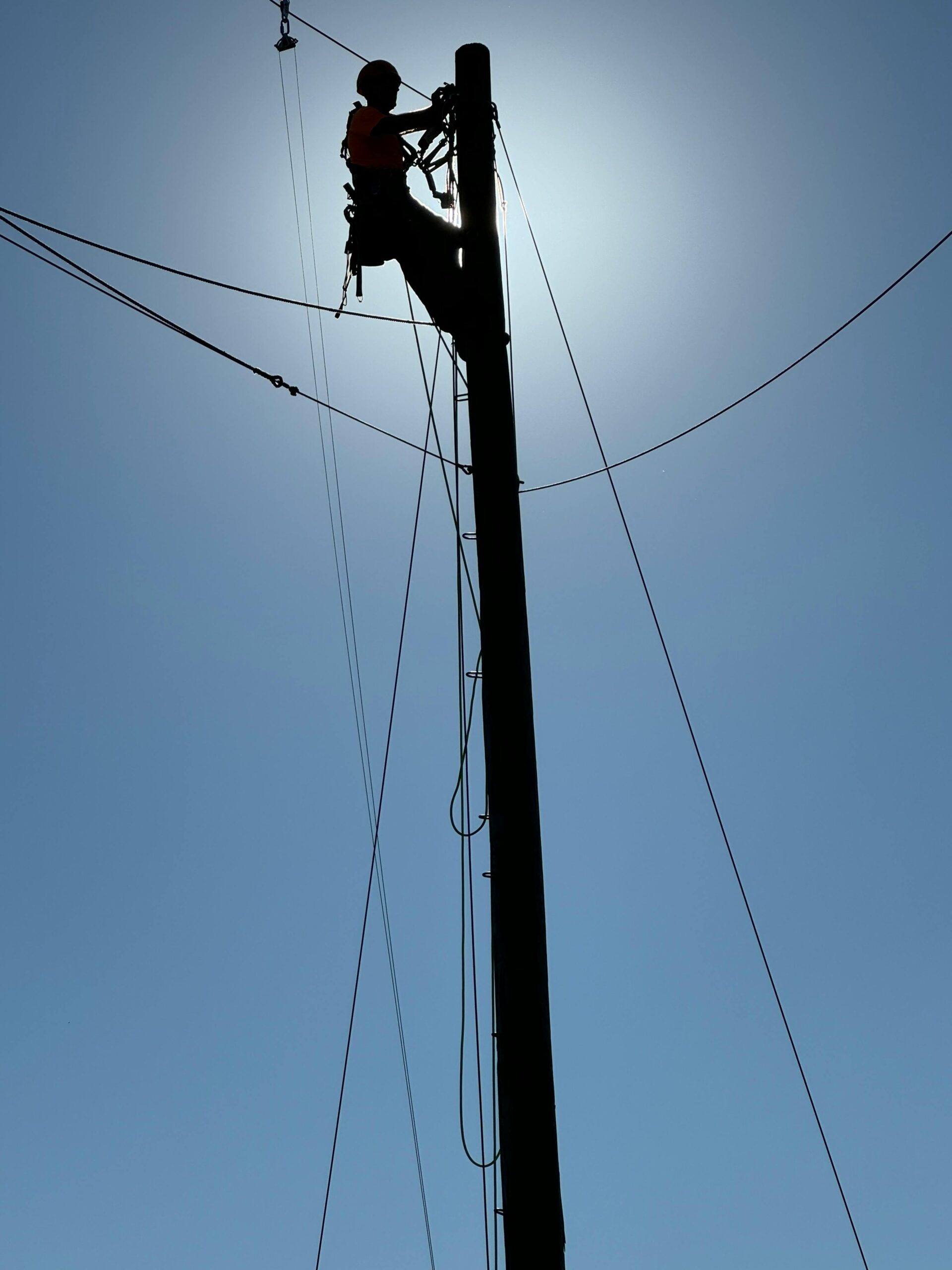 Lineman silhouetted against the sky while working on utility pole with cables.